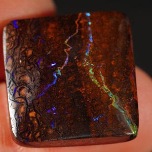 Könnte beinhalten: Ein quadratischer Boulder-Opal-Edelstein mit einer dunkelbraunen, texturierten Matrix und lebendigen, dünnen Adern aus schillerndem blauem und grünem Licht, die sich auf der Oberfläche spiegeln und natürliche Muster sowie eine glatte Politur zeigen.