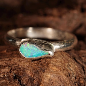 Könnte beinhalten: Ein Silberring mit einem einzelnen, tropfenförmigen Opal, der in einer glatten Metallfassung sitzt. Der Opal zeigt lebhafte grüne und blaue Schimmer, während der polierte Silberring ein abgerundetes, massives Profil aufweist.