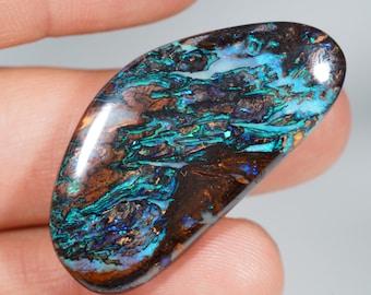 Puede incluir: Una gema pulida de forma ovalada con un patrón arremolinado de tonos marrones, turquesas y azules. La piedra tiene una superficie brillante y muestra un juego de color, con destellos de luz iridiscente. La gema se sostiene en una mano.