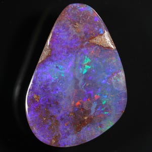 May include: A polished, teardrop-shaped opal gemstone. The stone displays a vibrant play-of-color with hues of purple, blue, green, and red. The opal has a dark base color with areas of brown and tan.