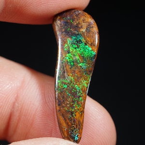 Könnte beinhalten: Ein polierter, länglicher Edelstein mit einer satten braunen Grundfarbe, akzentuiert durch leuchtende grüne und blaue irisierende Muster. Der Opal wird zwischen Fingern vor einem dunklen Hintergrund gehalten und zeigt seine natürliche Schönheit und einzigartigen Farbvariationen.