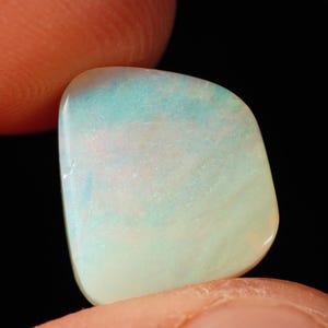 Könnte beinhalten: Ein polierter, unregelmäßiger, halbtransparenter Opal-Edelstein mit einer milchig-weißen Grundfarbe und irisierenden Blitzen in Hellblau und zartem Rosa auf seiner glatten, abgerundeten Oberfläche vor einem dunklen, schwarzen Hintergrund.