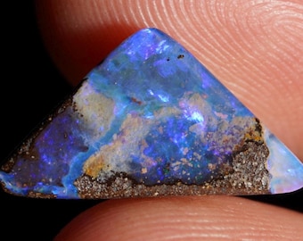 May include: A triangular-shaped opal gemstone with a vibrant display of blues, purples, and hints of yellow. The stone has a dark, textured base and a polished, iridescent surface, showcasing a natural, unique pattern.