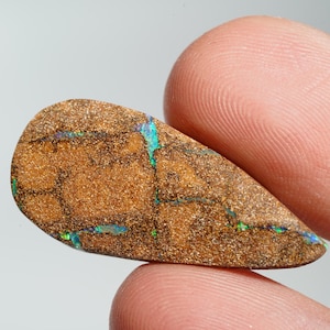 Könnte beinhalten: Ein brauner und goldener Opal-Edelstein mit grünen und blauen Farbschimmern. Der Opal ist tropfenförmig und hat eine raue, strukturierte Oberfläche.