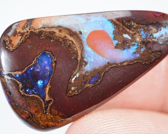 May include: A polished opal gemstone, shaped like a teardrop. The stone has a dark brown base with swirling patterns of orange, gold, blue, and turquoise. The opal's surface is smooth and reflective, displaying its natural colours and patterns.