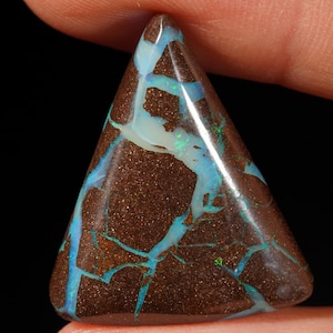 Pode incluir: Uma pedra preciosa de opala boulder de formato triangular com uma matriz de pedra de ferro castanho-escura e veios vibrantes de azul elétrico e verde que percorrem toda a superfície. A pedra tem um acabamento polido e liso que reflete a luz.