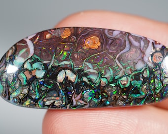 Puede incluir: Una gema pulida de forma ovalada con un patrón complejo de colores. La piedra presenta una base marrón oscuro con parches de naranja, morado y blanco. Vetas iridiscentes verdes y azules recorren la piedra, creando un diseño único y natural.