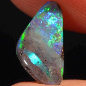 Könnte beinhalten: Ein polierter Opal-Edelstein mit dunkler Grundfarbe, der lebhafte Blitze von Grün, Blau und Lila zeigt. Der Edelstein hat eine glatte, abgerundete Form und eine glänzende Oberfläche, die sein inneres Farbspiel zeigt.