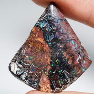 Puede incluir: Una gema con un patrón único de colores arremolinados, incluyendo marrón, negro, blanco y verde y azul iridiscente. La piedra se sostiene entre los dedos, mostrando su forma pulida en forma de diamante. La superficie de la gema tiene una apariencia texturizada.