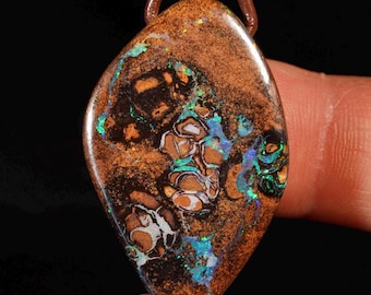 May include: A brown and black opal pendant with a leather cord. The gemstone features a unique pattern with hints of blue, green, and white. The pendant is a teardrop shape and is set against a black background.