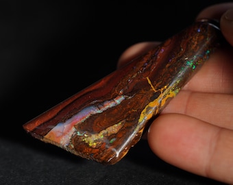 May include: A polished, elongated opal gemstone with a rich, earthy color palette. The stone displays a mix of deep reds, browns, and hints of blue, green, and yellow, with a unique, natural pattern. The opal is held in a hand.
