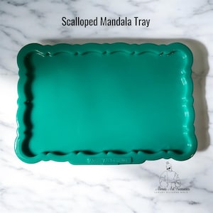 Pode incluir: Uma bandeja retangular turquesa com uma borda ondulada. A bandeja está rotulada como "Scalloped Mandala Tray" na parte superior e tem o texto "Almas Art Treasures" na parte inferior. A bandeja está sobre uma superfície de mármore branco.