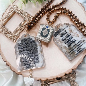 May include: Three gold-framed decorative pieces with Arabic calligraphy and a blue stone on a wooden surface. The pieces are hanging from a brown beaded necklace.