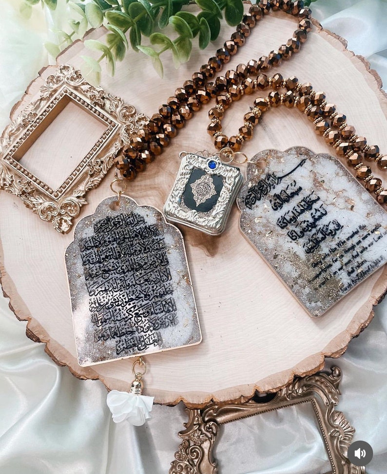 May include: Three gold-framed decorative pieces with Arabic calligraphy and a blue stone on a wooden surface. The pieces are hanging from a brown beaded necklace.