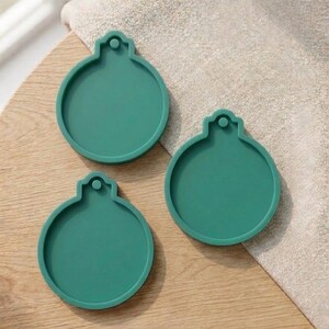 Può includere: Tre sottobicchieri a forma di ornamento color teal sono disposti su una superficie di legno chiaro. Ogni sottobicchiere ha una base circolare con un piccolo anello nella parte superiore. I sottobicchieri sono realizzati in un materiale solido.