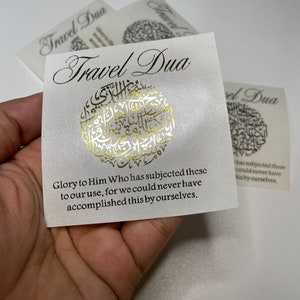Op de afbeelding: Een witte kaart met gouden letters die "Travel Dua" zegt en een Arabische inscriptie in goud bevat. De tekst onder de inscriptie luidt: "Eer aan Hem die deze dingen aan ons gebruik heeft onderworpen, want we zouden dit nooit zelf hebben kunnen bereiken."