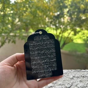 May include: A black rectangular pendant with an intricate white design of Arabic calligraphy. The pendant has a small loop at the top for attaching a chain.