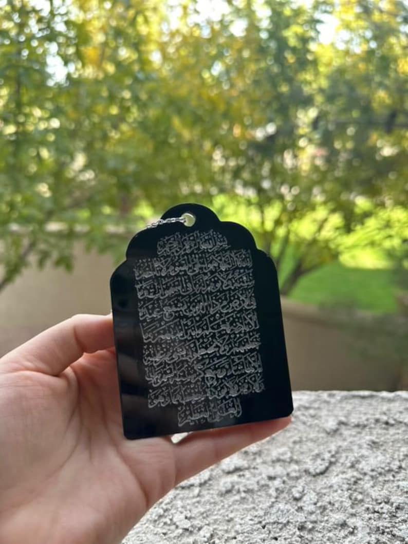 May include: A black rectangular pendant with an intricate white design of Arabic calligraphy. The pendant has a small loop at the top for attaching a chain.