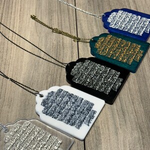 May include: Five different necklaces with pendants featuring Arabic calligraphy. The pendants are made of clear acrylic with different colored backgrounds: blue, green, black, white, and clear. Each pendant has a silver chain.