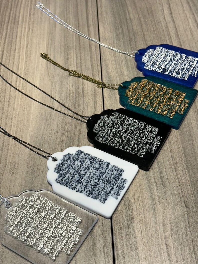 May include: Five different necklaces with pendants featuring Arabic calligraphy. The pendants are made of clear acrylic with different colored backgrounds: blue, green, black, white, and clear. Each pendant has a silver chain.