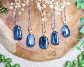 May include: Five blue kyanite stone pendants on silver chains. The gemstones have a polished, smooth surface and vary in shape. The pendants are displayed on a wooden surface with white flowers and green foliage.