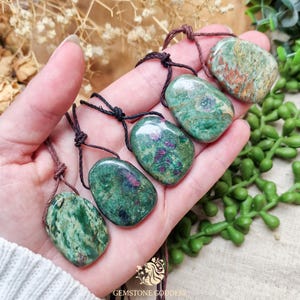 May include: Five polished, irregular-shaped green and red gemstone pendants, each suspended on a thin brown cord. The stones feature mottled patterns of deep forest green, lighter sage, and vibrant magenta-red inclusions throughout their surfaces.