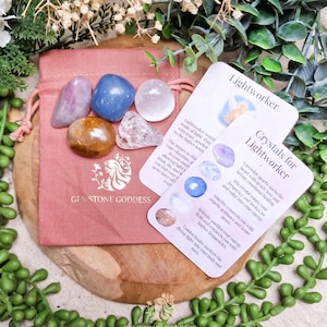 May include: A selection of colourful gemstones, including pink, blue, and clear stones, presented on a pink pouch. The pouch is branded with the text "GEMSTONE GODDESS" and a logo. Two cards with crystal information are also visible.