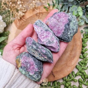 Könnte beinhalten: Vier rohe Rubinsteine mit einer Mischung aus rosa und grünen Farbtönen werden in einer Hand gehalten. Die Steine haben ein natürliches, unpoliertes Aussehen mit sichtbaren Texturen und Farbvariationen. Der Hintergrund umfasst eine Holzoberfläche und Grünpflanzen.