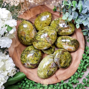 May include: A collection of green and white polished serpentine stones in a variety of shapes and sizes. The stones are arranged in a bowl on a wooden surface.