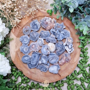 Puede incluir: Una colección de piedras preciosas de ágata pulidas y en bruto en tonos azules, grises y beige, dispuestas sobre una bandeja redonda de madera. Las piedras preciosas tienen patrones y texturas únicos. La bandeja está rodeada de vegetación y flores blancas.