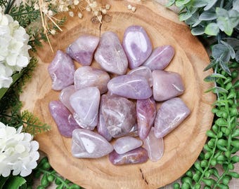 May include: A collection of polished amethyst gemstones in varying shades of purple and lavender, displayed on a rustic wooden plate. The gemstones are smooth and rounded, with natural variations in color and texture. The image is surrounded by greenery and white flowers.