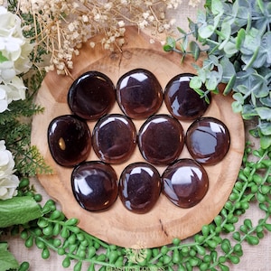 Pode incluir: Uma coleção de dez pedras lisas, de cor marrom escura, polidas e arredondadas. As pedras estão dispostas em um padrão circular sobre uma superfície de madeira.