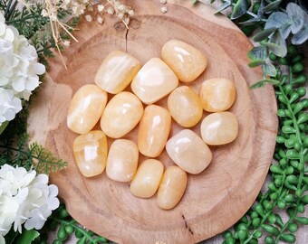 May include: A collection of smooth, polished orange calcite stones arranged on a round wooden bowl. The stones vary in size and shape, with a warm, translucent glow. The bowl is surrounded by white flowers and green foliage.