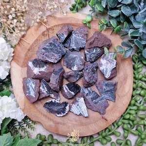 May include: A collection of rough, raw hematite stones, showcasing a deep reddish-brown hue with striking silver and black metallic inclusions. The stones are arranged on a wooden plate, surrounded by greenery and white flowers.