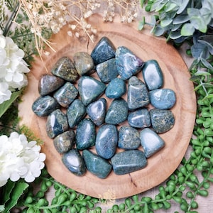 May include: A collection of polished, tumbled gemstones in shades of teal, green, and blue, arranged on a round wooden plate. The stones vary in size and shape, with a smooth, glossy finish. The plate is surrounded by greenery and white flowers.