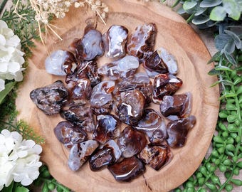 Puede incluir: Una colección de piedras de ágata pulidas en tonos marrón, blanco y gris, dispuestas sobre una placa de madera redonda. Las piedras tienen una textura suave y brillante y patrones variados. El arreglo está rodeado de vegetación y flores blancas.
