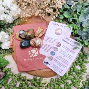 Può includere: Una collezione di pietre preziose lucide in vari colori, tra cui marrone, verde e crema, esposte su un sacchetto rosa. Sono presenti anche due carte con testo sui cristalli. Il sacchetto riporta la scritta "GEMSTONE GODDESS".