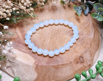 Könnte beinhalten: Ein hellblaues Perlenarmband, bestehend aus runden, undurchsichtigen Perlen. Das Armband wird auf einer Holzoberfläche präsentiert, umgeben von Grün und Trockenblumen. Die Perlen haben ein weiches, milchiges Aussehen.