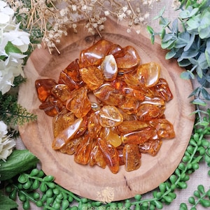 May include: A wooden bowl filled with polished amber gemstones. The amber stones vary in shape and size, displaying shades of orange and brown. The bowl sits on a surface surrounded by greenery and white flowers, creating a natural aesthetic.
