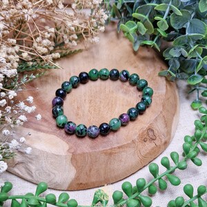 Pode incluir: Uma pulseira de contas com pedras preciosas redondas em tons de verde, preto e roxo-avermelhado. A pulseira é exibida em uma tábua de madeira, rodeada por vegetação e flores secas. As pedras preciosas têm aproximadamente 0,8 cm de diâmetro.