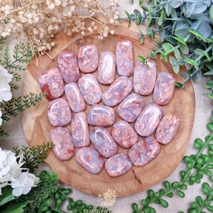 Puede incluir: Una colección de piedras preciosas pulidas en tonos rosa, lavanda y rojo, dispuestas sobre una tabla de madera. Las piedras tienen un aspecto veteado y están rodeadas de vegetación y flores secas. La imagen incluye el texto "GEMSTONE GODDESS".