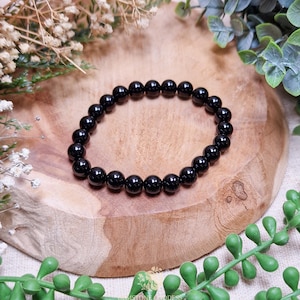 Könnte beinhalten: Ein schwarzes Perlenarmband mit polierten, runden Perlen. Das Armband wird auf einer Holzoberfläche präsentiert, umgeben von Grün und weißen Blumen. Die Perlen haben einen Durchmesser von etwa 8 mm.
