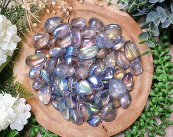 May include: A selection of iridescent aura quartz tumbled stones in shades of blue, purple, and gold, presented on a round wooden bowl. The stones are smooth and polished, reflecting light. The bowl is surrounded by greenery and white flowers.