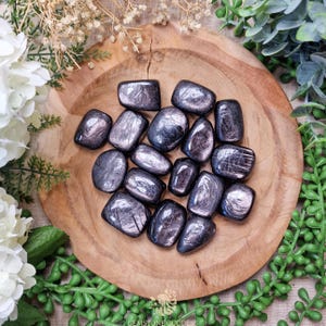 Könnte beinhalten: Eine Sammlung von geschliffenen schwarzen Obsidian-Steinen mit irisierendem Schimmer. Die Steine sind in einer Holzschale angeordnet.