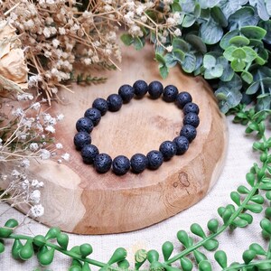 Op de afbeelding: Een zwarte lavasteen kralenarmband. De armband is gemaakt van ronde, poreuze kralen. De armband wordt gepresenteerd op een houten oppervlak, omringd door groen en droogbloemen.