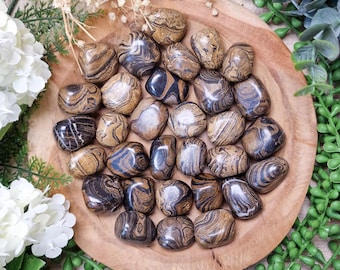May include: A collection of polished, tumbled stones in shades of brown, black, and cream, arranged on a wooden plate. The stones have unique, swirling patterns. The image is framed with white flowers and green foliage, creating a natural aesthetic.