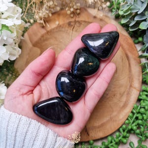May include: Four smooth, polished black obsidian tumbled stones with iridescent green and gold flecks. The stones are arranged in a row on a hand.