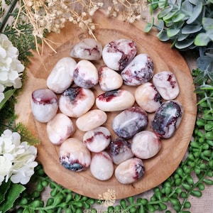 Op de afbeelding: Een verzameling gepolijste edelstenen in wit, crème en bordeaux, gerangschikt op een houten bord. De stenen variëren in grootte en vorm, sommige vertonen donkerrode en groene insluitsels. De afbeelding is omgeven door groen en witte bloemen.