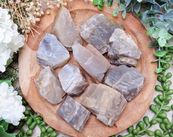 May include: A collection of raw, uncut moonstone crystals in varying shapes and sizes. The stones display a range of neutral colors, including gray, beige, and hints of blue. They are arranged on a round, wooden plate, surrounded by greenery and white flowers.