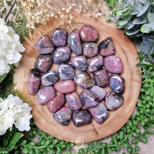 Puede incluir: Una colección de piedras de rodonita pulidas en tonos rosa, gris y negro, dispuestas sobre una placa de madera. Las piedras varían en tamaño y forma, con un acabado liso y brillante. La disposición está rodeada de vegetación y flores blancas.